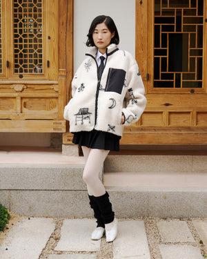 A woman with short dark hair and a neutral expression stands before a wooden building, wearing a white Sundae School Kiwa Fleece Zip-Up with black patterns, a black skirt, white tights, black leg warmers, and white shoes.