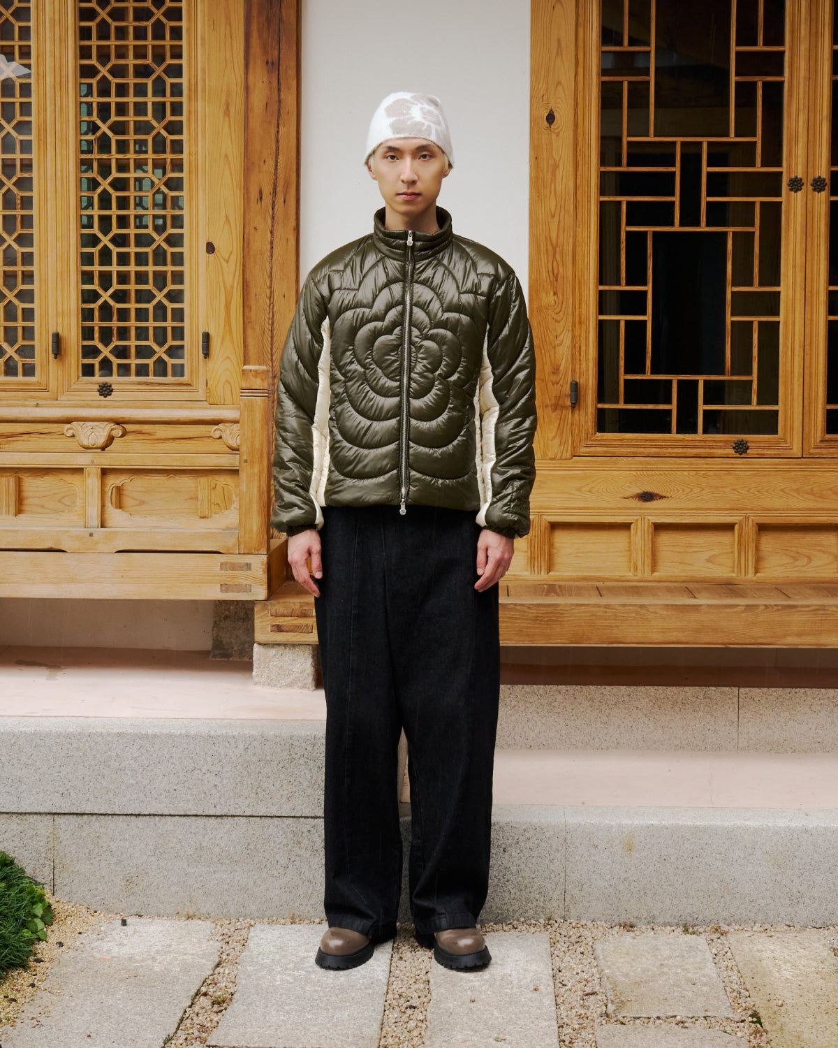 A man stands in front of a building wearing the Sundae School OLIVE OATMEAL MOCHI SPIRAL PUFFER jacket.