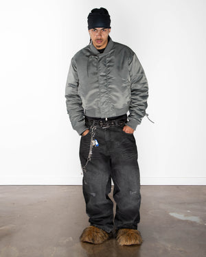 A person stands indoors against a white wall, wearing the Sundae School Khaki Flower Channeled Bomber with adjustable flower sleeves, loose black jeans, a black beanie, chain accessories, and brown furry shoes on a stained concrete floor.