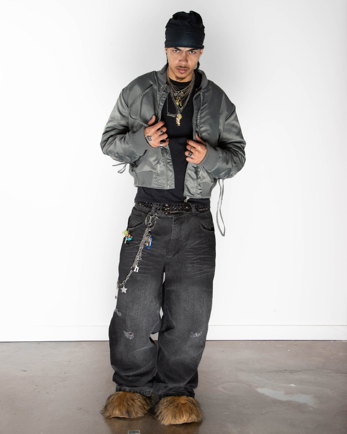 A person stands against a plain white wall wearing the Sundae School KHAKI FLOWER CHANNELED BOMBER with adjustable flower sleeves, a black shirt, baggy distressed jeans, a black beanie, layered necklaces, and brown furry shoes. They look at the camera and hold their jacket open.
