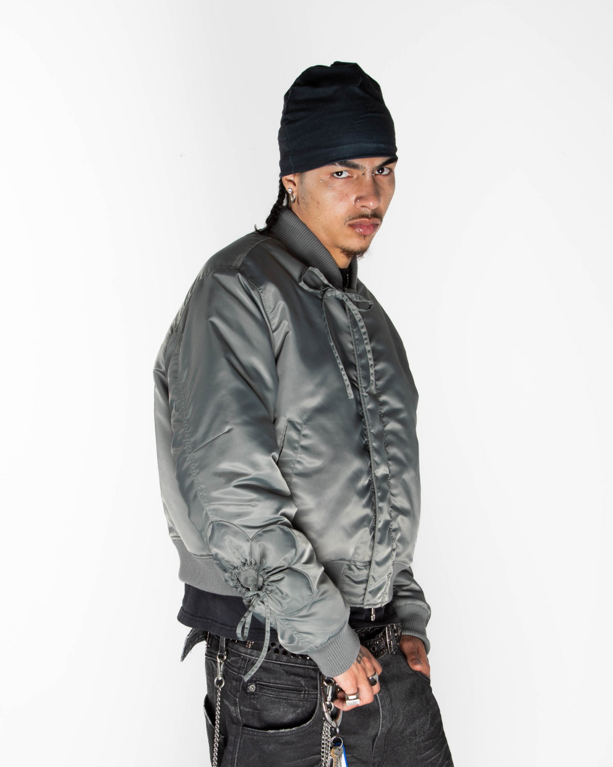 A person wearing a khaki Sundae School Flower Channeled Bomber with adjustable flower sleeves, a black beanie, and dark pants stands against a white background, gazing seriously at the camera with their hands by their sides.