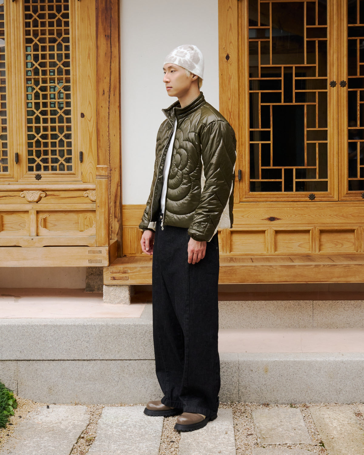 A man in the Sundae School OLIVE OATMEAL MOCHI SPIRAL PUFFER, black wide-leg pants, brown shoes, and a white patterned beanie stands sideways before a traditional wooden building with geometric windows.