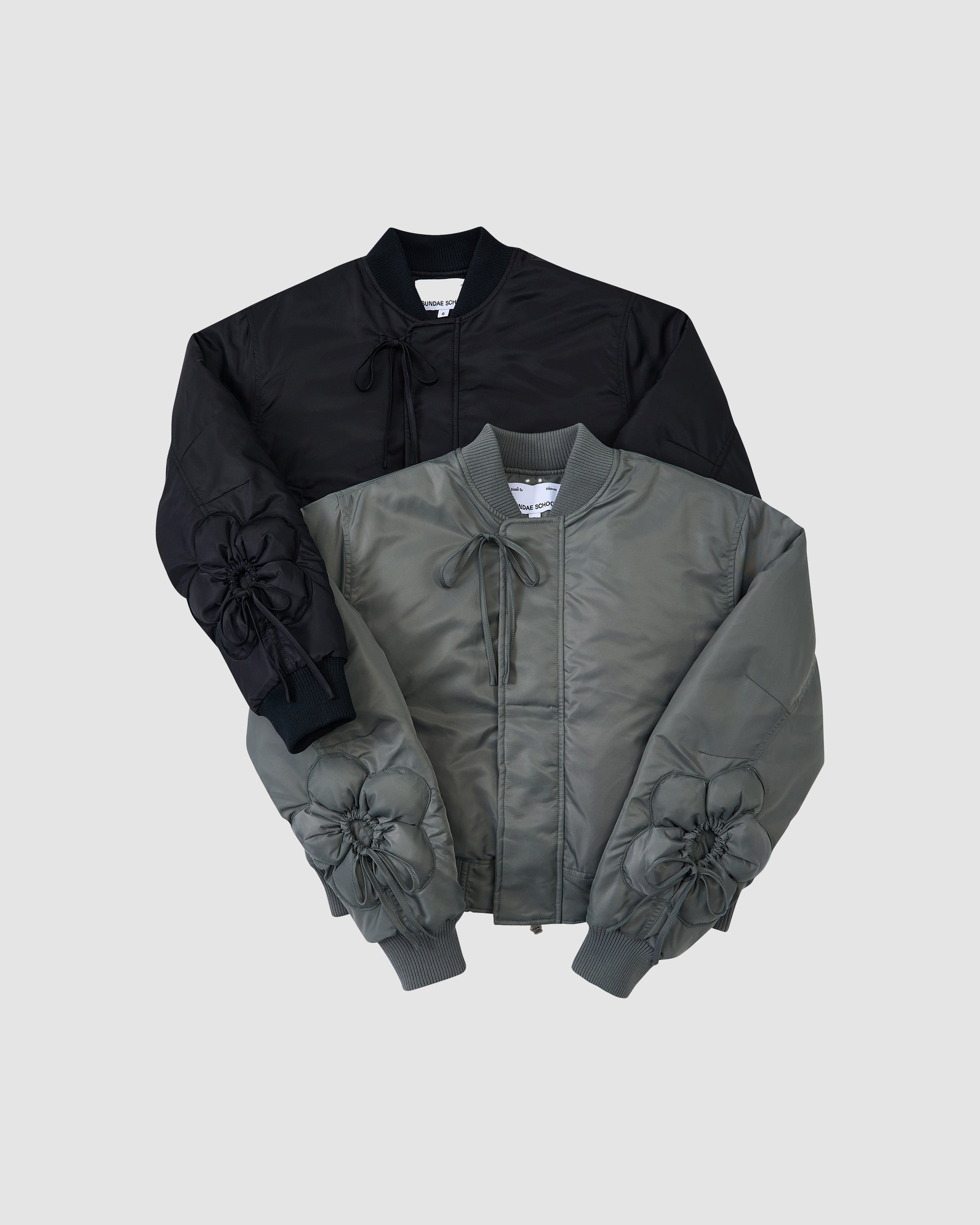 The BLACK FLOWER CHANNELED BOMBER by Sundae School, shown in black and olive green on a light gray background, features ruched sleeves with drawstrings plus ribbed cuffs and hems.