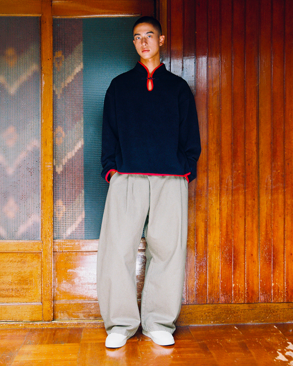 A person wearing a dark blue long-sleeved shirt with red trim, loose-fitting light grey STONE KIEF HANBOK CHINO PANTS, and white sneakers.