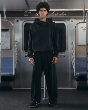 A person with curly hair stands in the middle of an empty subway car, facing the camera and wearing Sundae School's BLACK WASH HANBOK HOODIE—featuring an oversized hood—paired with black pants and black shoes.