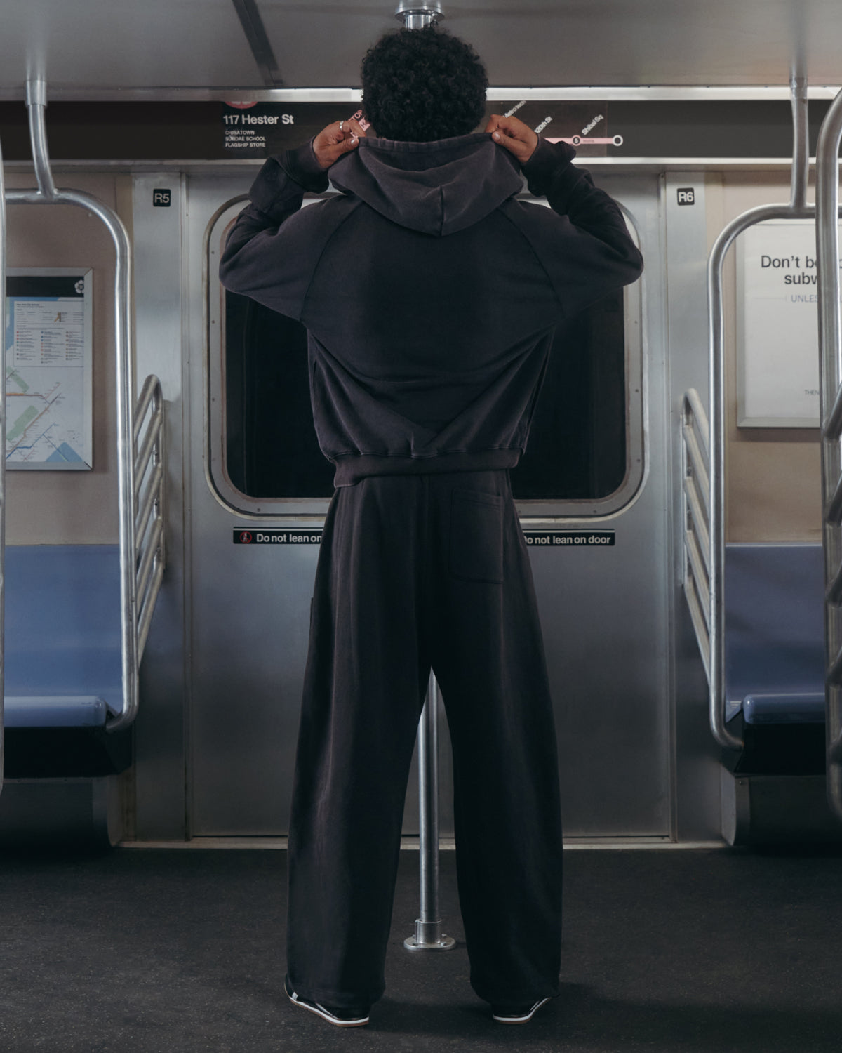 A person wearing the Sundae School BLACK WASH HANBOK HOODIE stands on a subway train with their back to the camera, holding the hood with both hands. Subway doors and signs appear in the background.