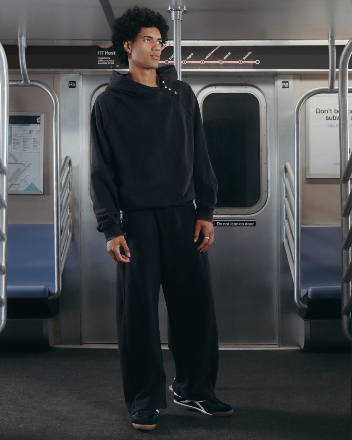 A person wearing the Sundae School BLACK WASH HANBOK HOODIE with matching loose-fit pants stands alone in an empty subway car, gazing off to the side. The relaxed cotton sweatshirt, featuring an oversized hood, echoes the quiet ambiance of the scene, with rows of subway seats and metal poles visible in the background.