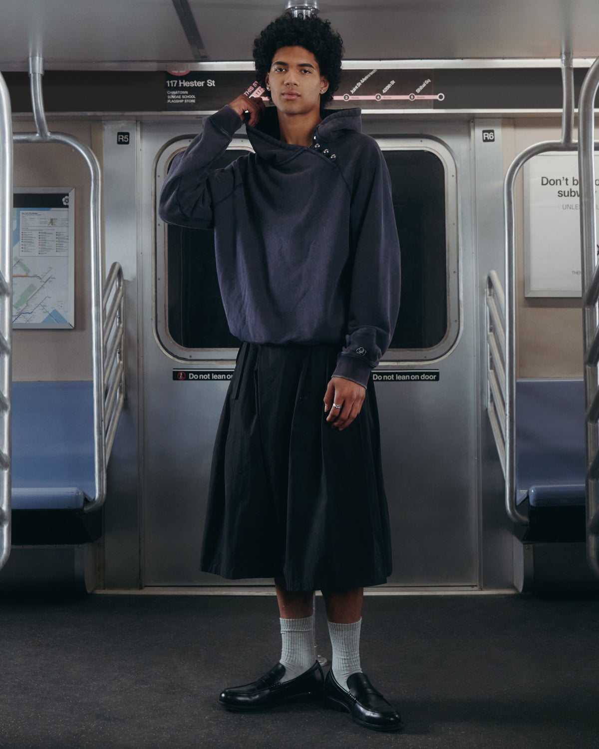 A person stands in an empty subway car, wearing Sundae School’s NAVY WASH HANBOK HOODIE with its oversized hood, paired with a black skirt, white socks, and black shoes. Their expression is thoughtful as they lightly touch their neck with one hand.