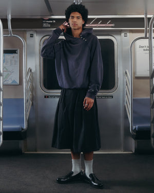 A person stands in an empty subway car, wearing Sundae School’s NAVY WASH HANBOK HOODIE with its oversized hood, paired with a black skirt, white socks, and black shoes. Their expression is thoughtful as they lightly touch their neck with one hand.