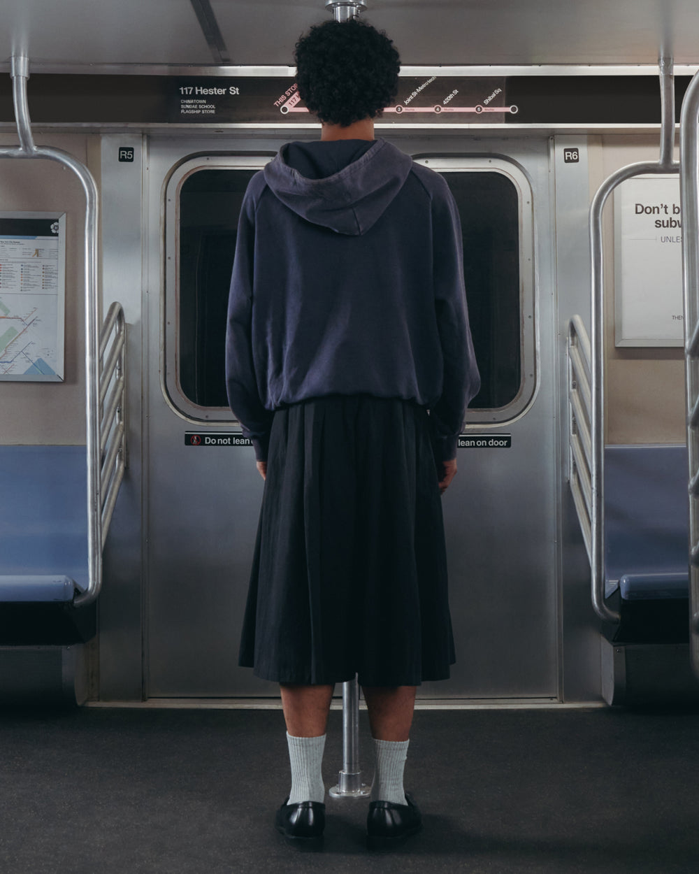 A person dressed in a dark hoodie, socks, and the Sundae School ASYMMETRIC CHIMA CULOTTE stands facing the subway doors inside a mostly empty subway car. A subway map and safety signs are visible above the door.
