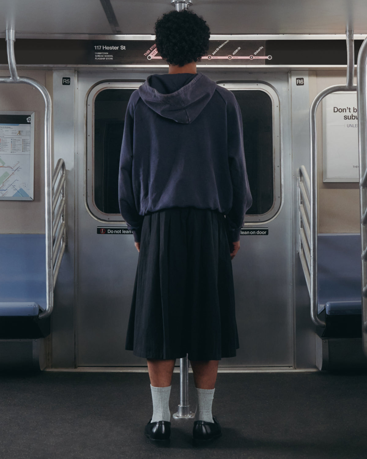 A person with short curly hair, dressed in the Sundae School NAVY WASH HANBOK HOODIE featuring an oversized hood, along with a long skirt, white socks, and black shoes, stands facing the subway doors in an empty subway car.