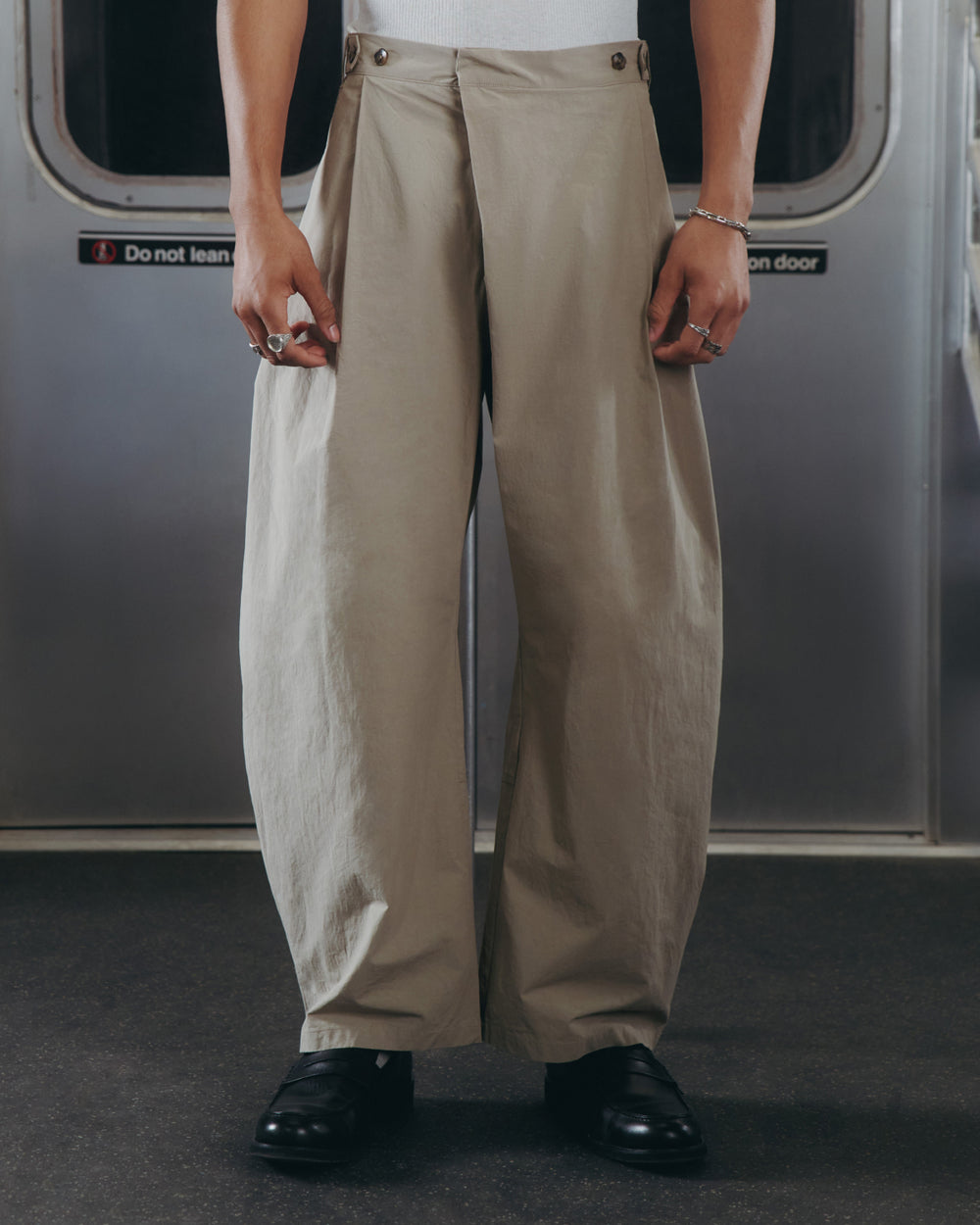 A person stands in a subway car wearing Sundae School WHEAT HANBOK LINEN PANTS with an adjustable waist, a white top, black shoes, and silver rings and bracelets. Subway doors and a warning sign are visible in the background.