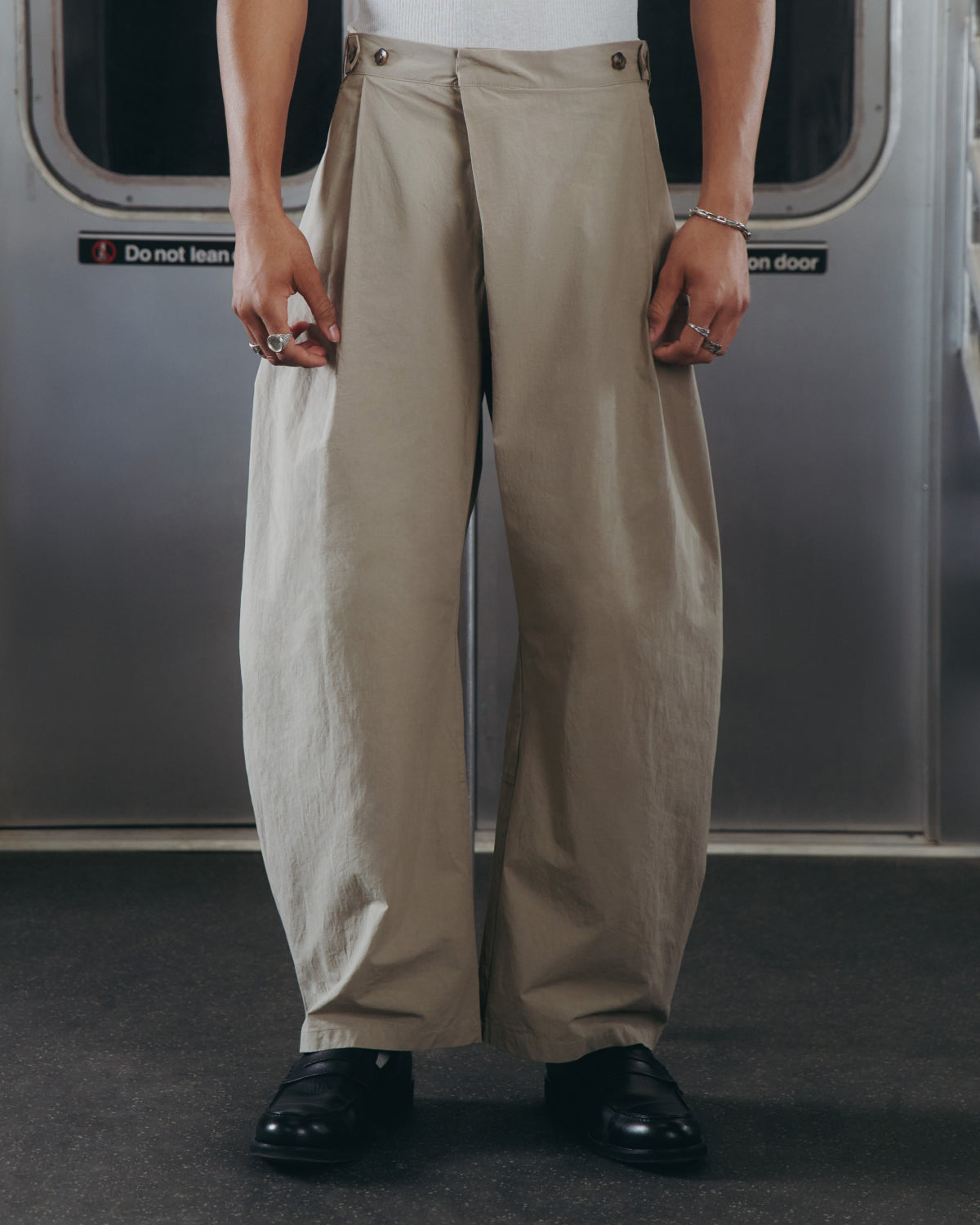 A person stands in a subway car wearing Sundae School WHEAT HANBOK LINEN PANTS with an adjustable waist, a white top, black shoes, and silver rings and bracelets. Subway doors and a warning sign are visible in the background.