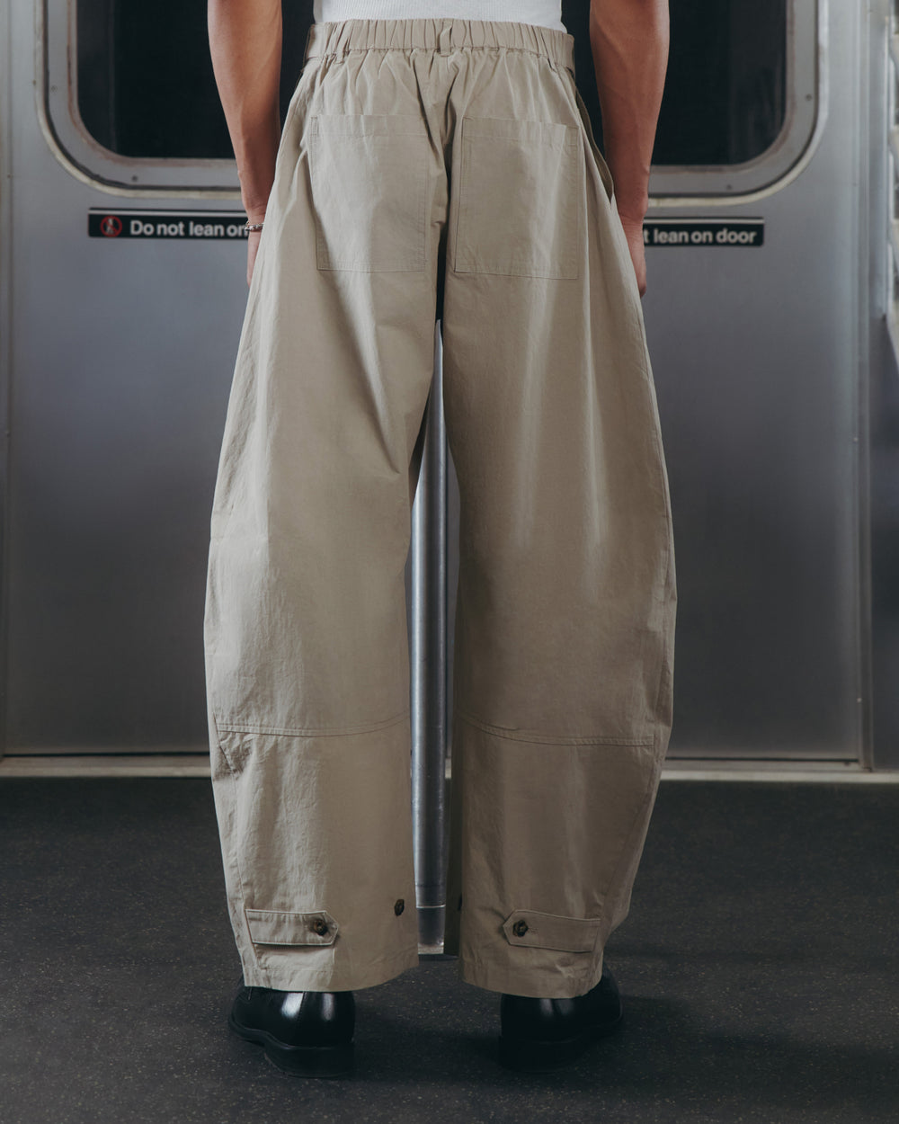 A person stands in a subway car wearing Sundae School’s WHEAT HANBOK LINEN PANTS, known for their wide barrel leg silhouette, adjustable waist, and ankle button details; the photo captures only their lower half from behind.