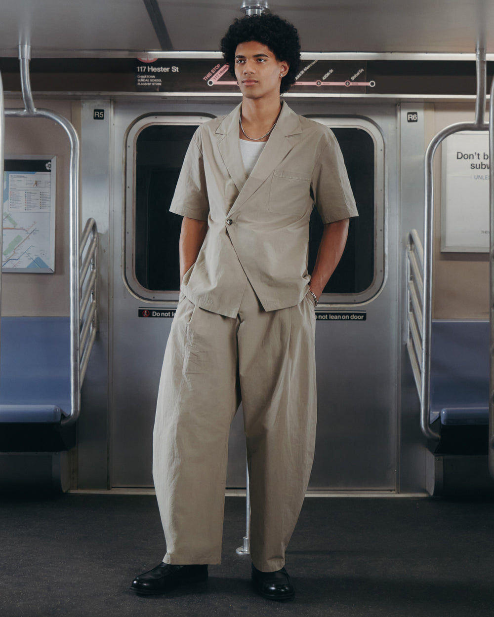 A person with curly hair stands in a subway car wearing Sundae School's WHEAT HANBOK LINEN PANTS, paired with a loose light beige short-sleeve suit and black shoes, hands in pockets, gazing slightly to the side.
