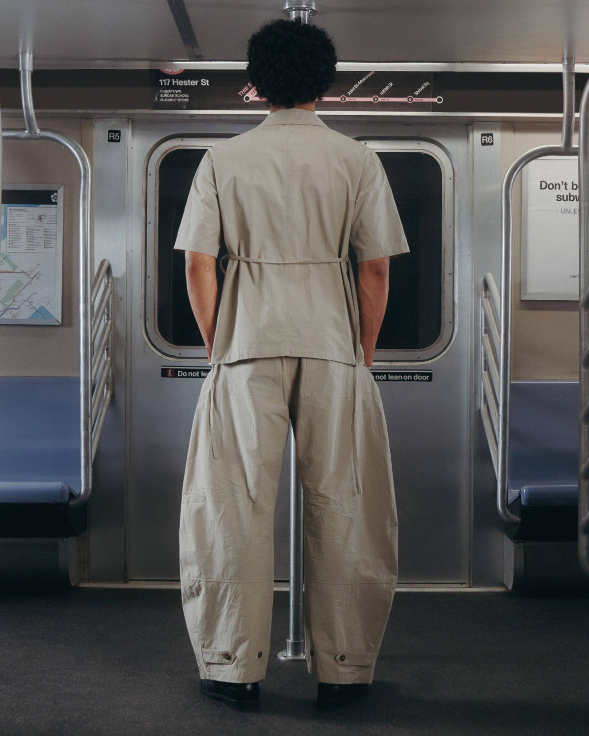 A person with curly hair, wearing the Sundae School WHEAT HANBOK LINEN SHIRT, stands facing closed subway doors in an empty car with blue seats and metal handrails.