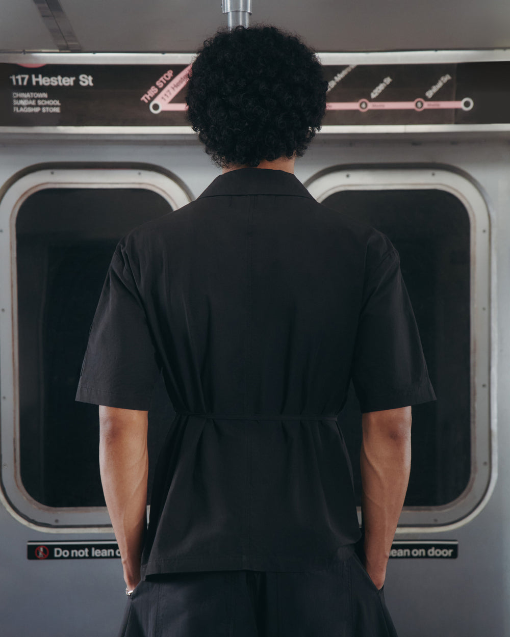 A person with short curly hair, wearing a Sundae School BLACK HANBOK LINEN SHIRT, stands with their back to the camera inside a subway train beneath a station sign that reads 117 Hester St.