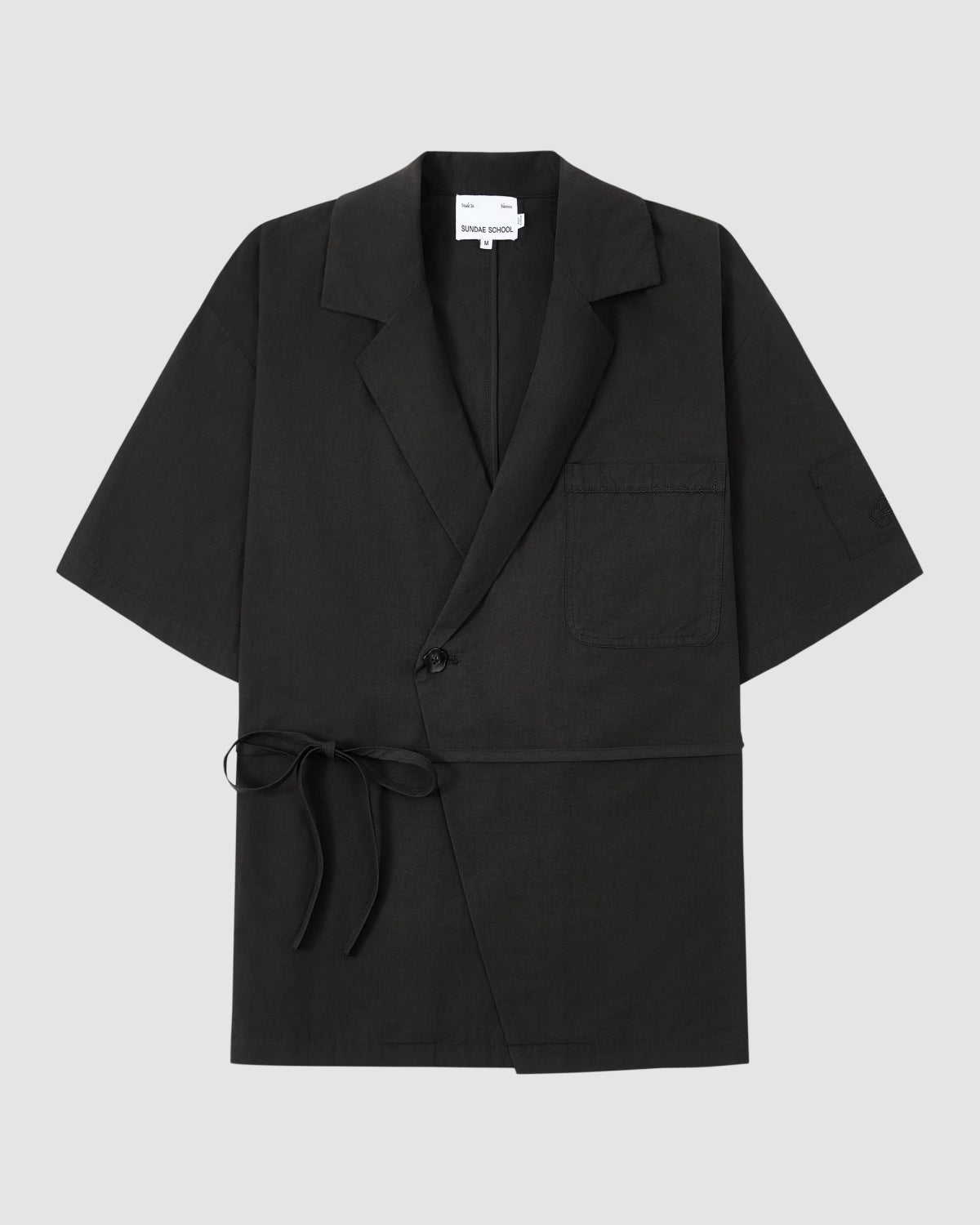 The BLACK HANBOK LINEN SHIRT by Sundae School is a black, short-sleeve cotton-linen blend with a notched collar, chest pocket, single button closure, and side tie—styled like a modern hanbok shirt on a light grey background.