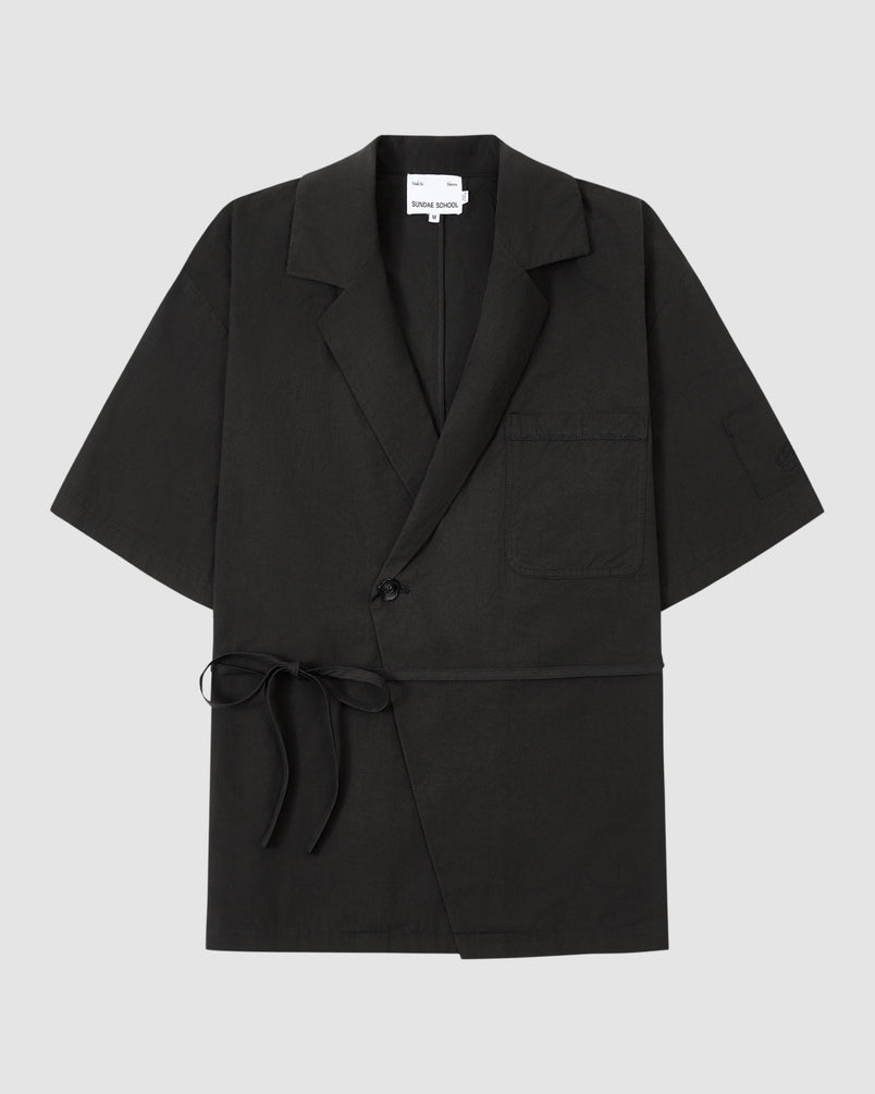 The BLACK HANBOK LINEN SHIRT by Sundae School is a black, short-sleeve cotton-linen blend with a notched collar, chest pocket, single button closure, and side tie—styled like a modern hanbok shirt on a light grey background.