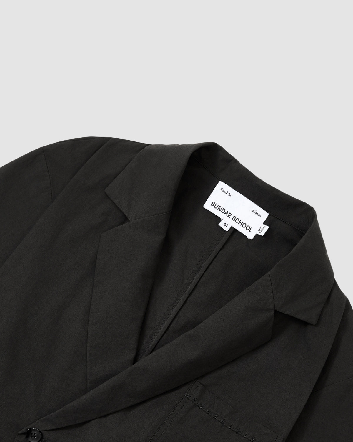 Close-up of the BLACK HANBOK LINEN SHIRT by Sundae School, featuring a notched collar and cotton-linen blend, with a visible white brand tag at the inner neck, laid flat on a light gray background.