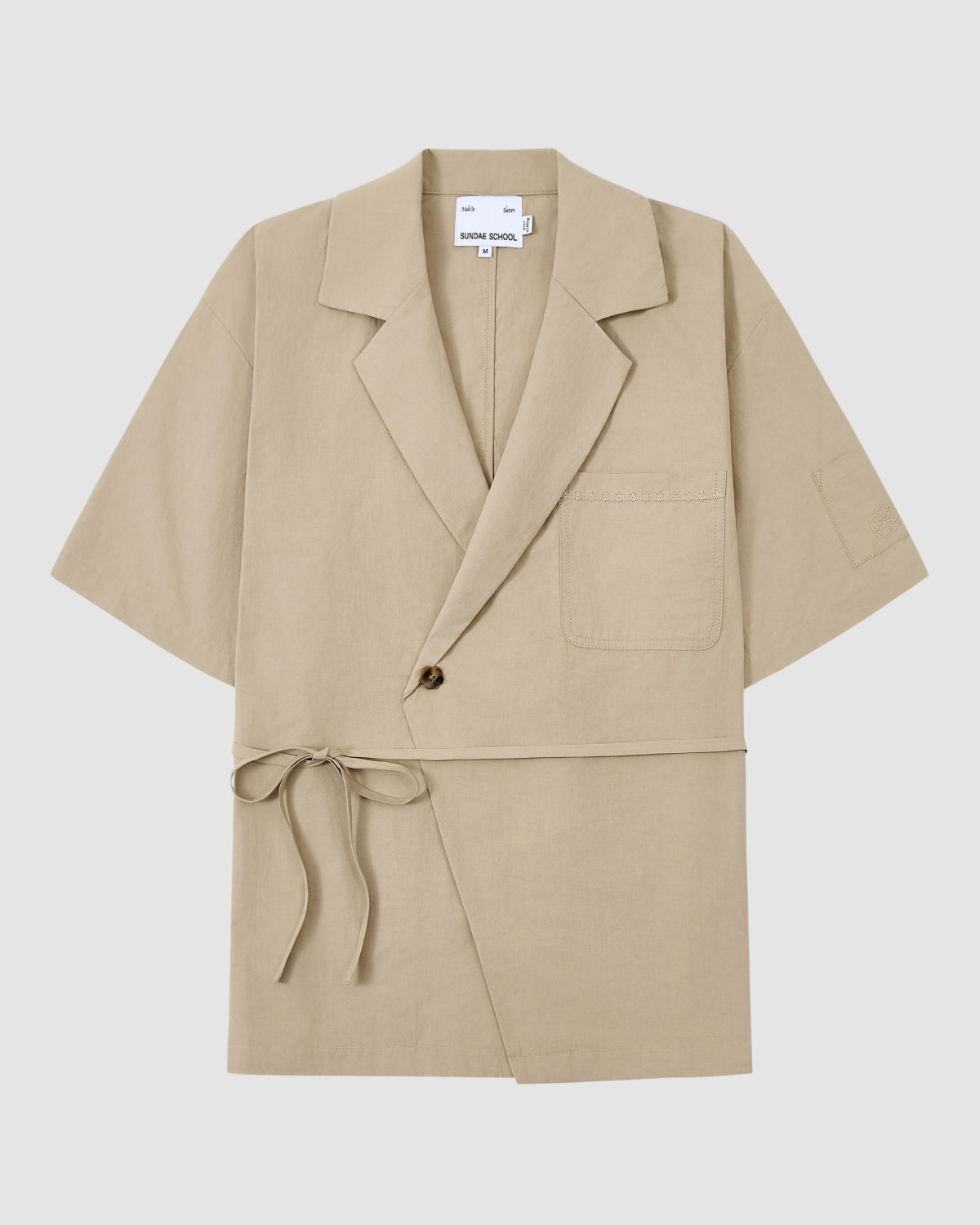The WHEAT HANBOK LINEN SHIRT by Sundae School is a beige short-sleeve wrap jacket made from breathable cotton-linen blend, with a notched lapel, chest pocket, single-button closure, and side tie at the waist. Shown on a plain white background.