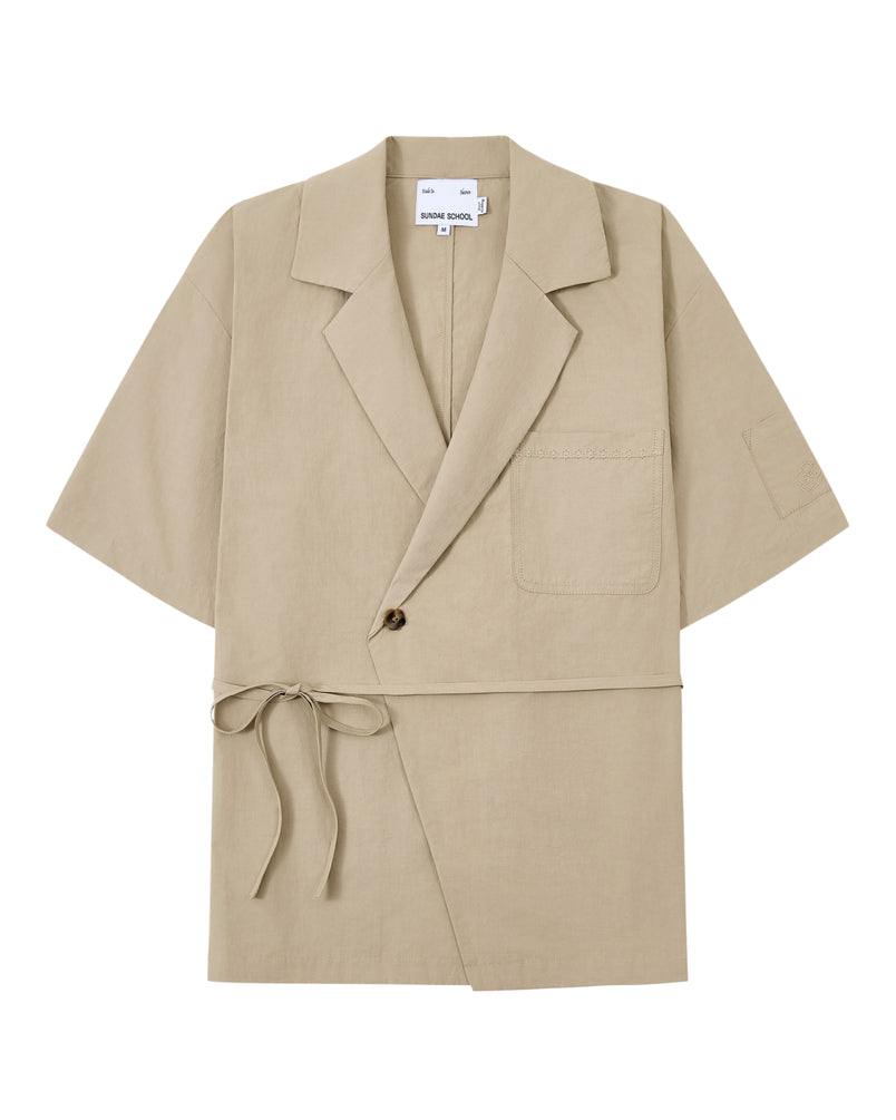 The WHEAT HANBOK LINEN SHIRT by Sundae School is a beige short-sleeve jacket made from a breathable cotton-linen blend, featuring a notched collar, single chest pocket, one-button closure, and side tie detail inspired by the modern hanbok shirt, shown laid flat on a white background.