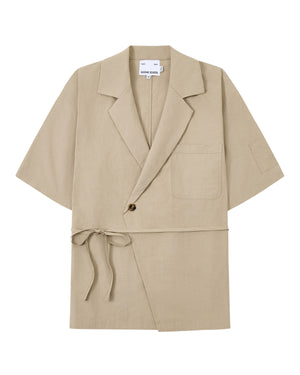 The WHEAT HANBOK LINEN SHIRT by Sundae School is a beige short-sleeve jacket made from a breathable cotton-linen blend, featuring a notched collar, single chest pocket, one-button closure, and side tie detail inspired by the modern hanbok shirt, shown laid flat on a white background.