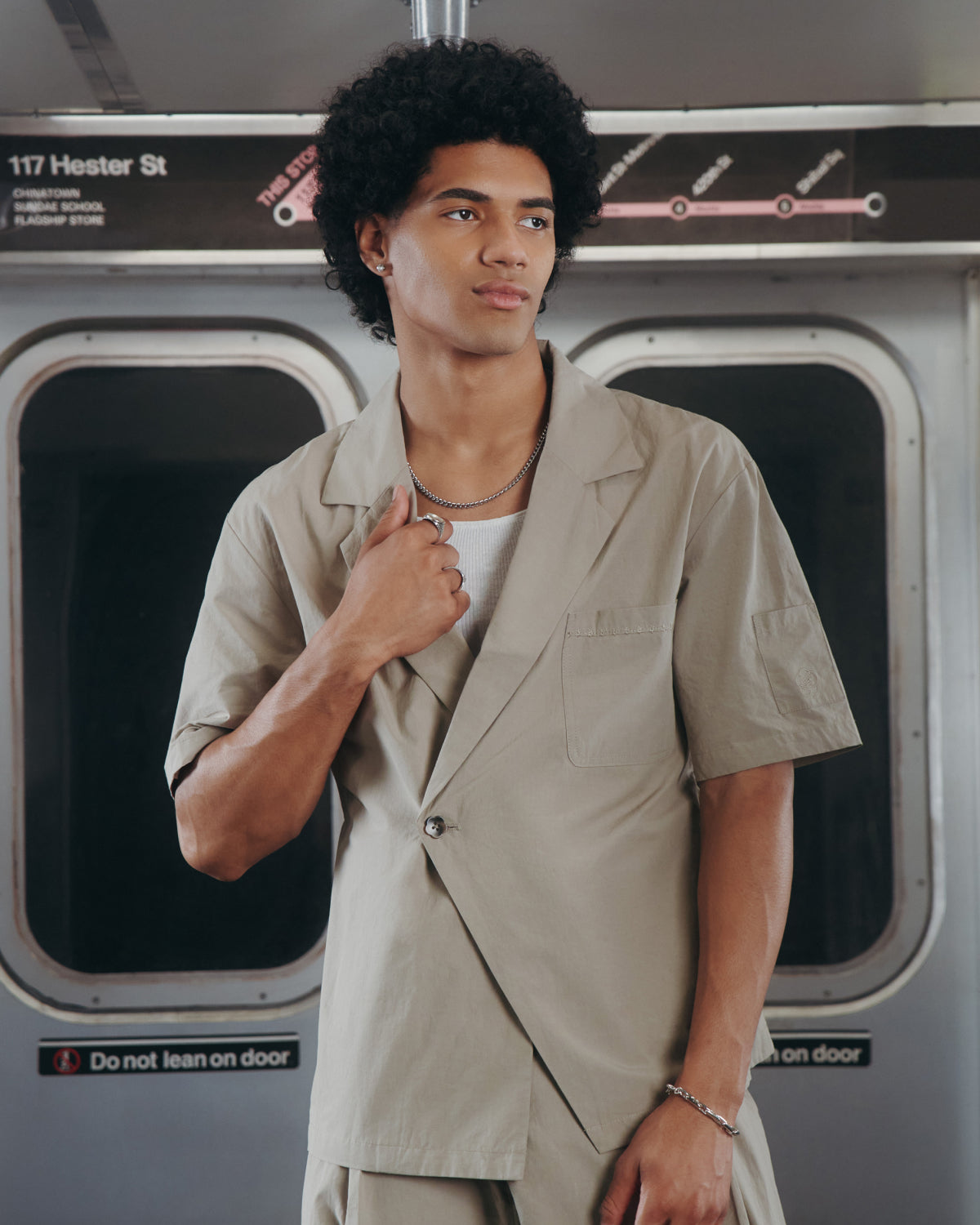 A person with curly hair, wearing the Sundae School WHEAT HANBOK LINEN SHIRT, stands in a subway car holding the lapel. A subway map and a sign reading 