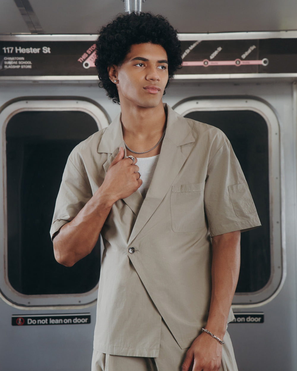 A person with curly hair, wearing the Sundae School WHEAT HANBOK LINEN SHIRT, stands in a subway car holding the lapel. A subway map and a sign reading "117 Hester St" are visible above the train doors.