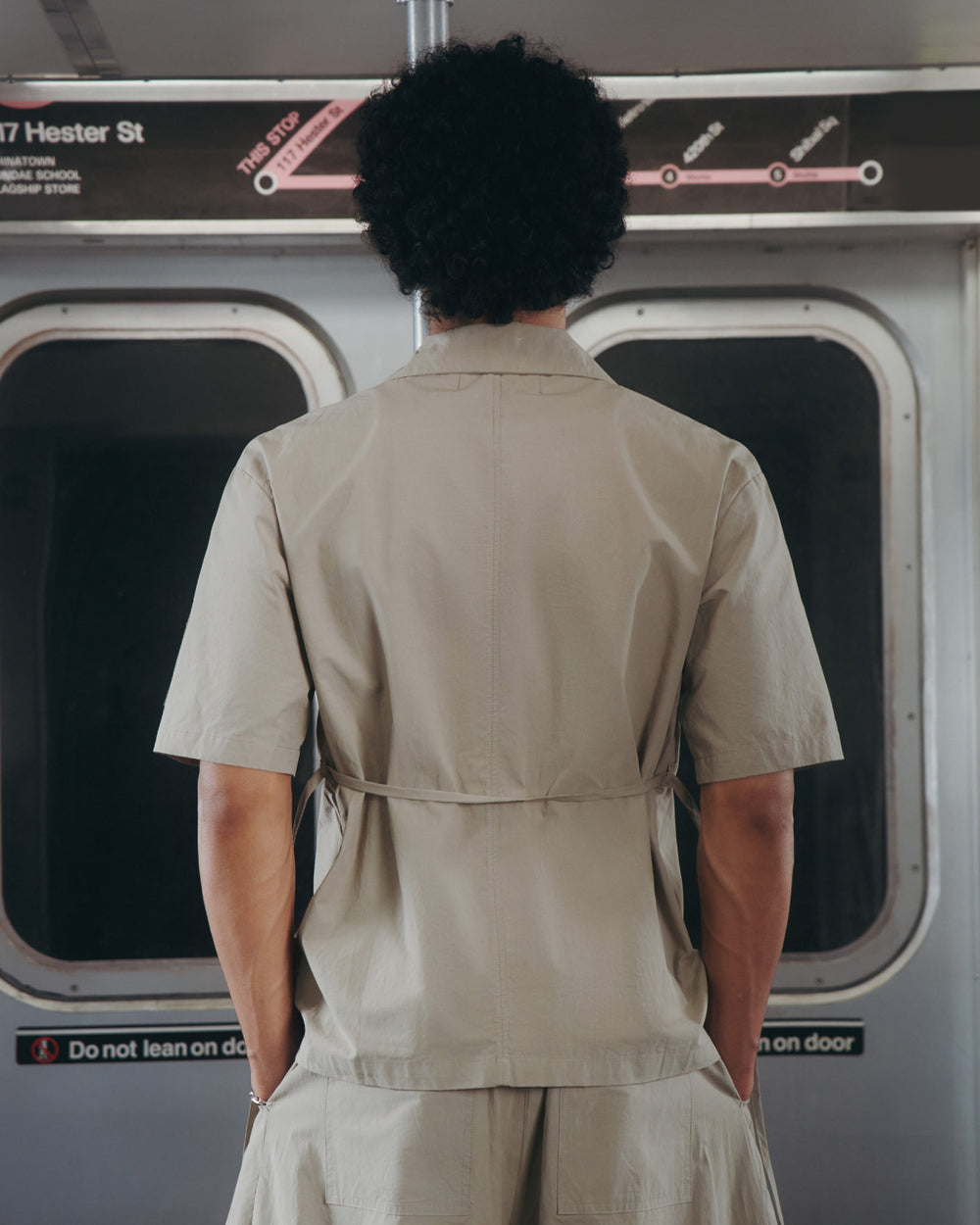 Seen from behind on a subway, a person with curly hair wears the Sundae School WHEAT HANBOK LINEN SHIRT—crafted from a cotton-linen blend—in light beige. Subway map and doors are in the background.