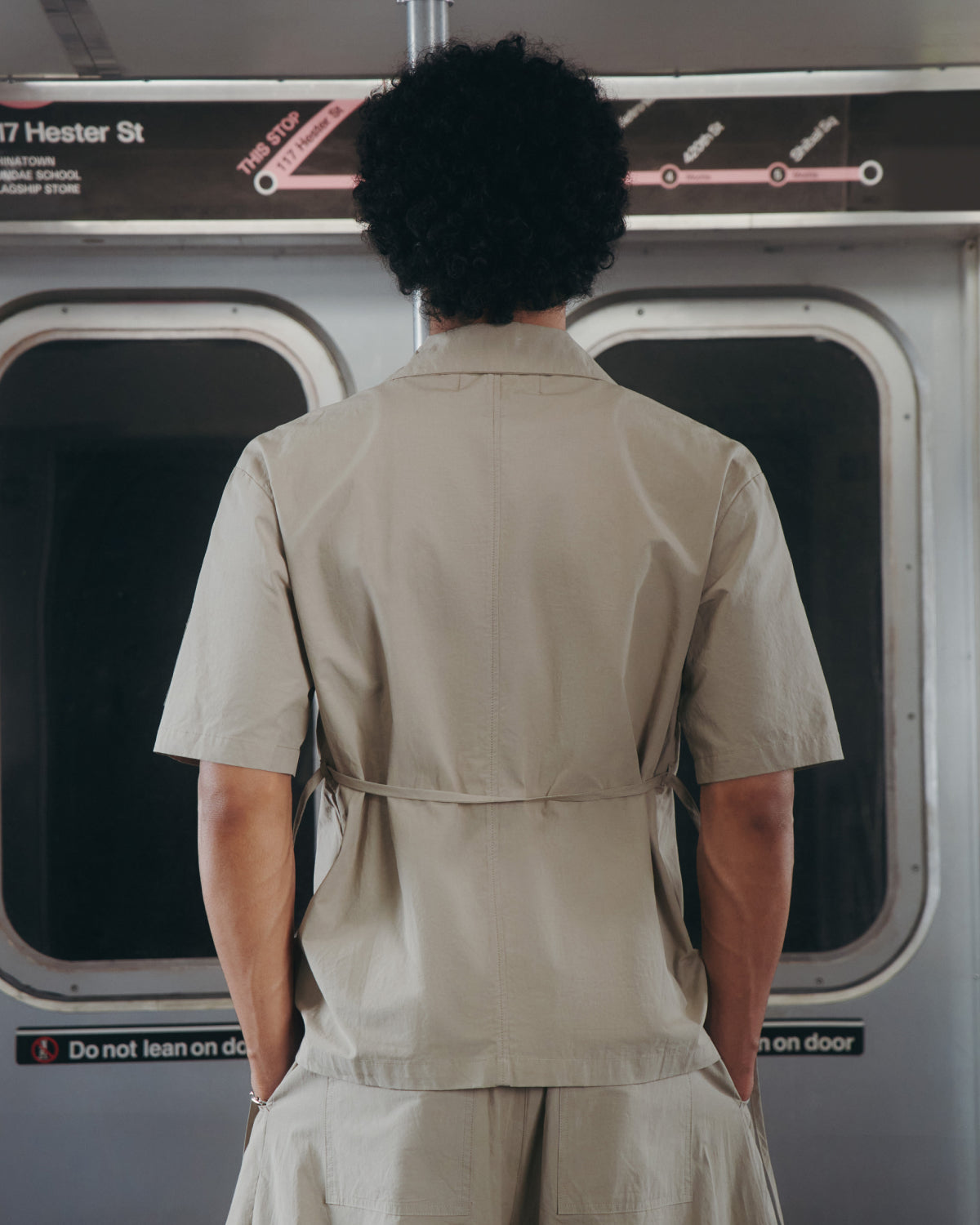 Seen from behind on a subway, a person with curly hair wears the Sundae School WHEAT HANBOK LINEN SHIRT—crafted from a cotton-linen blend—in light beige. Subway map and doors are in the background.