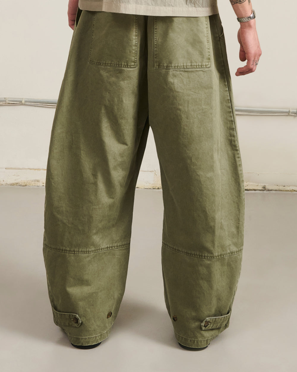 A person wearing very baggy olive green cargo pants, shown from the back against a plain off-white studio wall.