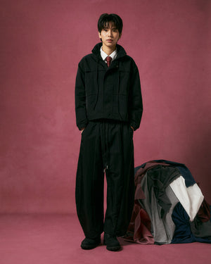 A person stands against a mauve backdrop wearing the BLACK HANBOK TRACK JACKET by Sundae School, paired with loose black pants, black shoes, a white shirt, and a red tie. Colorful folded and draped fabrics are arranged to their right.