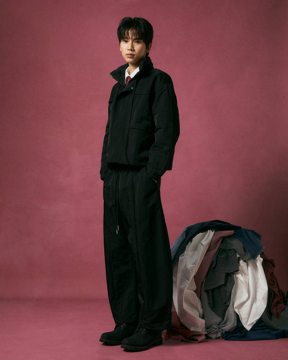 A person wearing Sundae School’s BLACK HANBOK BALLOON TRACK PANTS stands on a pink background next to draped, multicolored fabric, looking at the camera with a neutral expression.