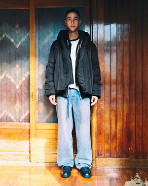 A person stands indoors in front of wooden doors, wearing the Sundae School BLACK HEAVYWEIGHT MOCHI PUFFER over a white t-shirt, paired with loose light blue jeans and black shoes. The floor is shiny and the background features patterned glass.
