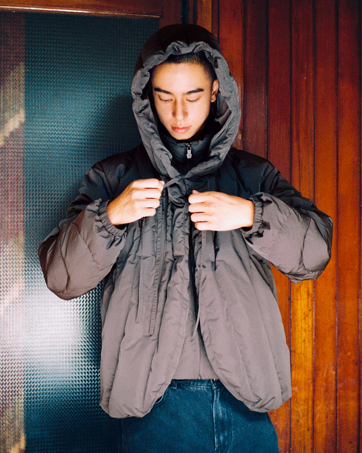 A person wearing the Sundae School CHARCOAL HEAVYWEIGHT MOCHI PUFFER, a hooded gray jacket with duck down fill, stands indoors against a wooden wall, looking down while adjusting the drawstrings of their jacket.
