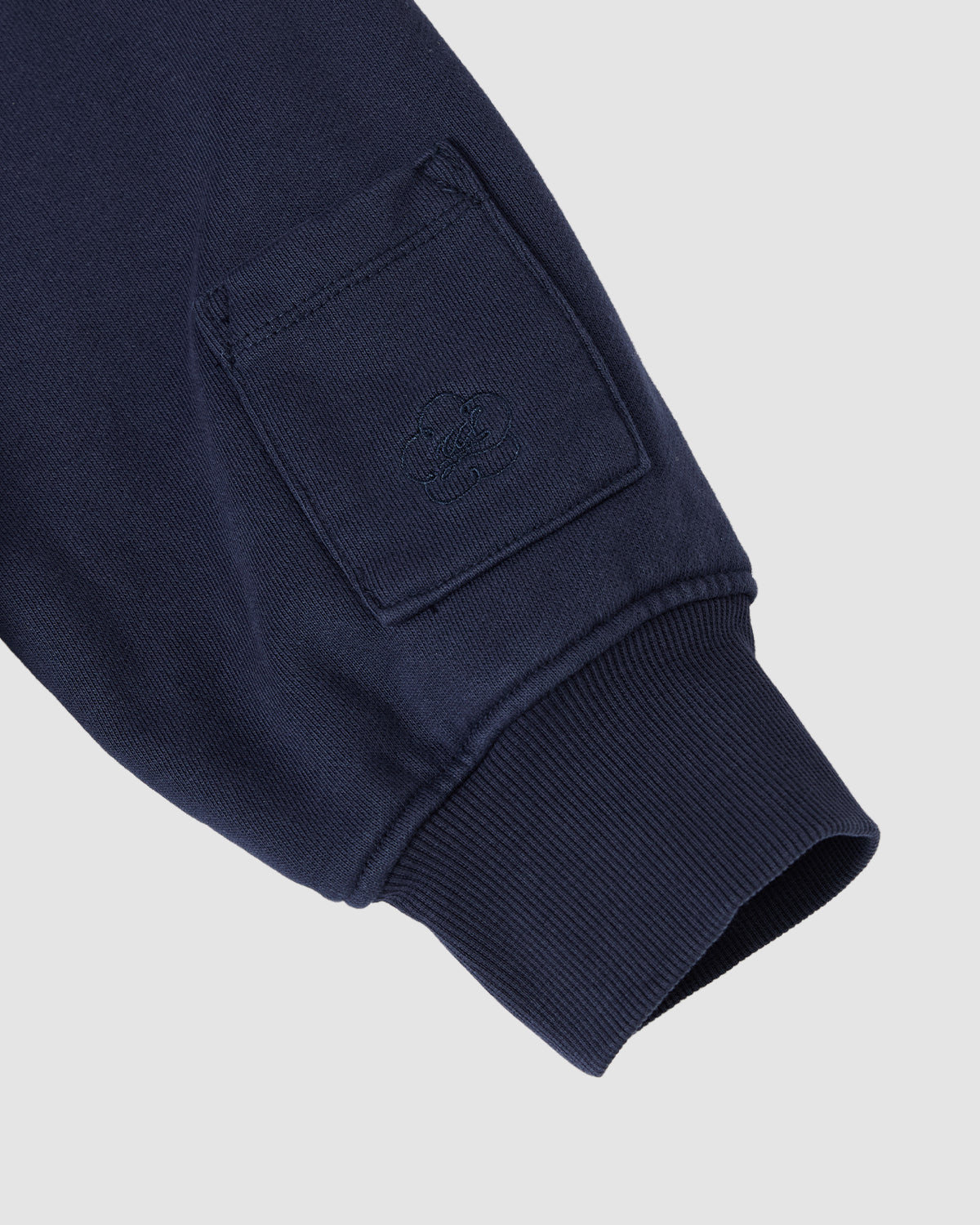 Close-up of the Sundae School NAVY HESTER QUARTER ZIP’s navy cotton sleeve, showcasing a ribbed cuff and small stash pocket with embossed logo on the upper arm, set against a light gray background.