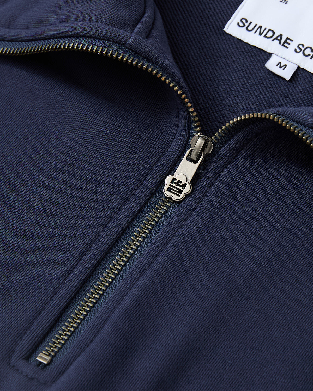 Close-up of the NAVY HESTER QUARTER ZIP by Sundae School, an oversized navy sweater with a metallic quarter zip partially open. The interior collar shows a white label reading SUNDAE SCHOOL M.