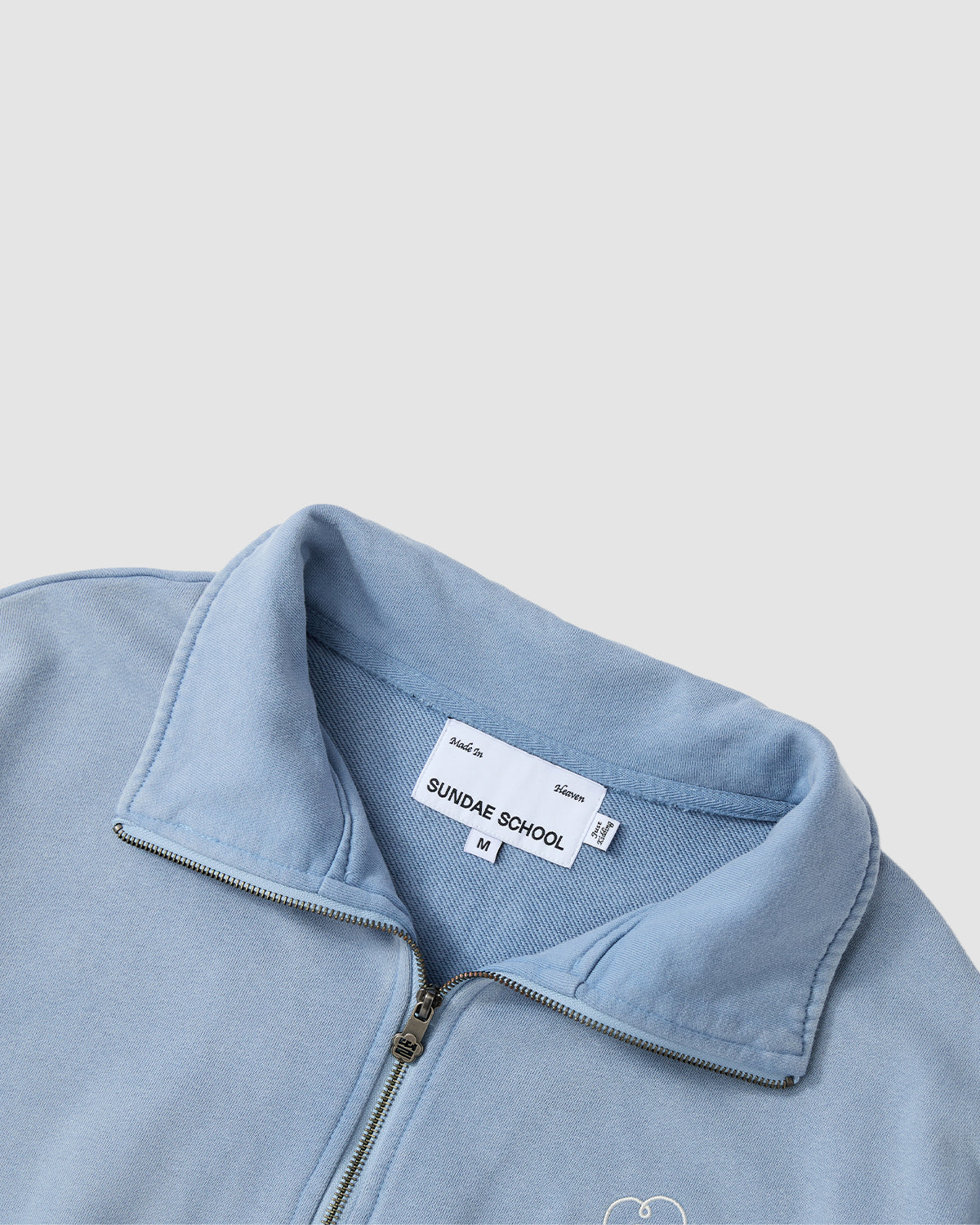 The SKY BLUE HESTER QUARTER ZIP by Sundae School is a light blue fleece jacket with a stand-up collar, hidden stash pocket, minimal stitching, and a simple design. The inner tag shows Sundae School branding and size M.