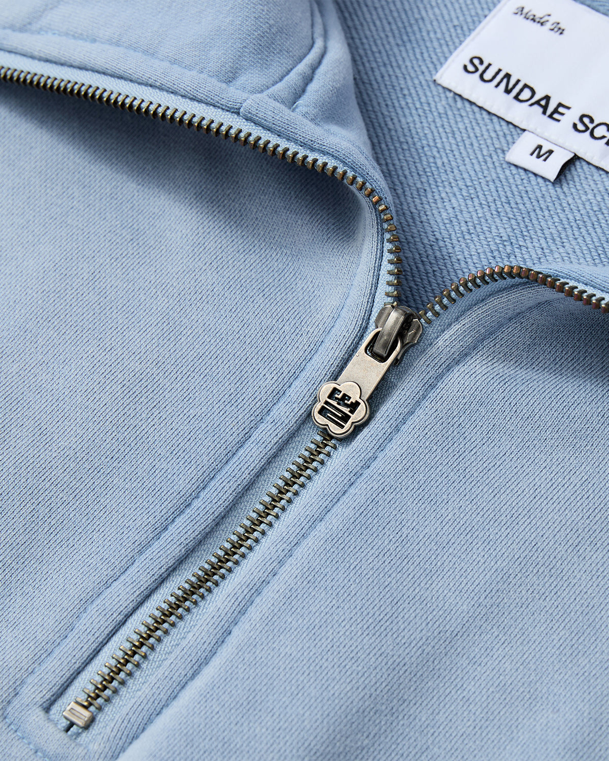 Close-up of the SKY BLUE HESTER QUARTER ZIP by Sundae School, partially unzipped to display its metallic zipper. A white label inside shows the brand and M for medium. The soft, textured fabric is light blue and oversized.