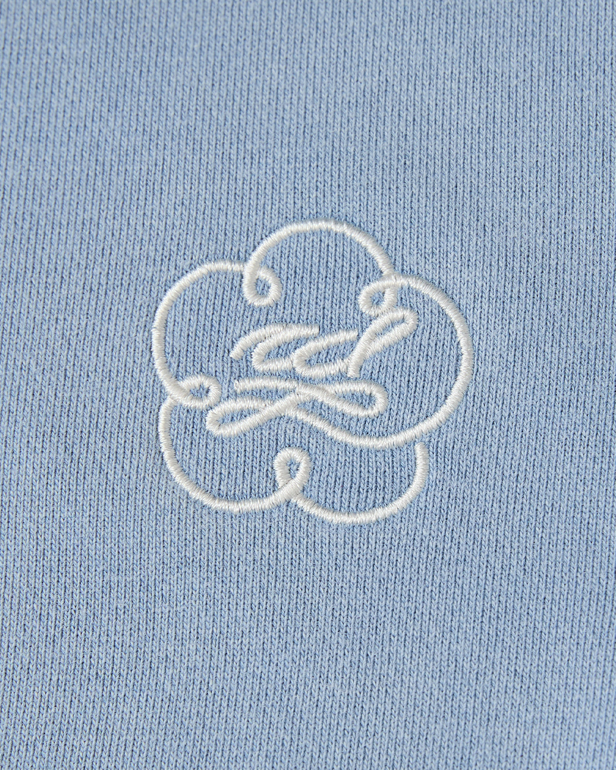 A close-up of the Sundae School SKY BLUE HESTER QUARTER ZIP, a light blue cotton pullover with a white embroidered abstract flower-like design and curved lines.
