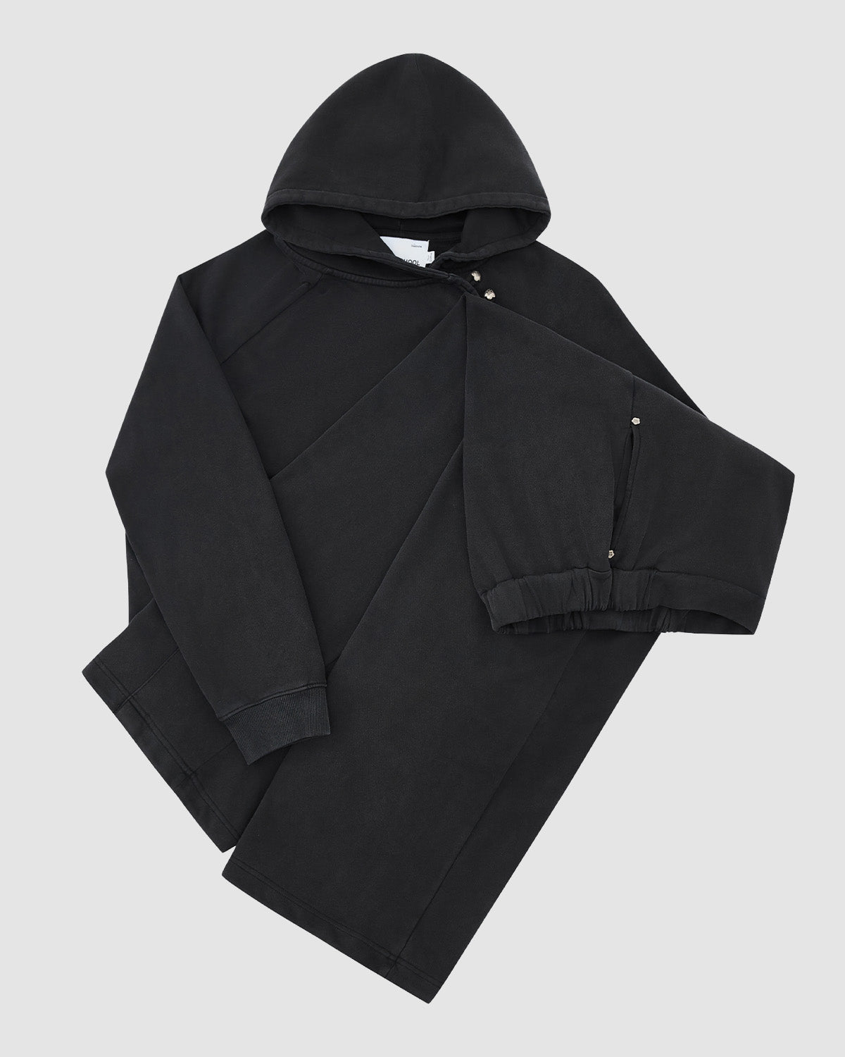 The FADE BLACK WRAP SWEATBAJI by Sundae School, a black hooded jacket with long sleeves, snap buttons, and elastic cuffs, features Hanbok-inspired details. It's laid flat on a light gray background with the left sleeve folded over the front.