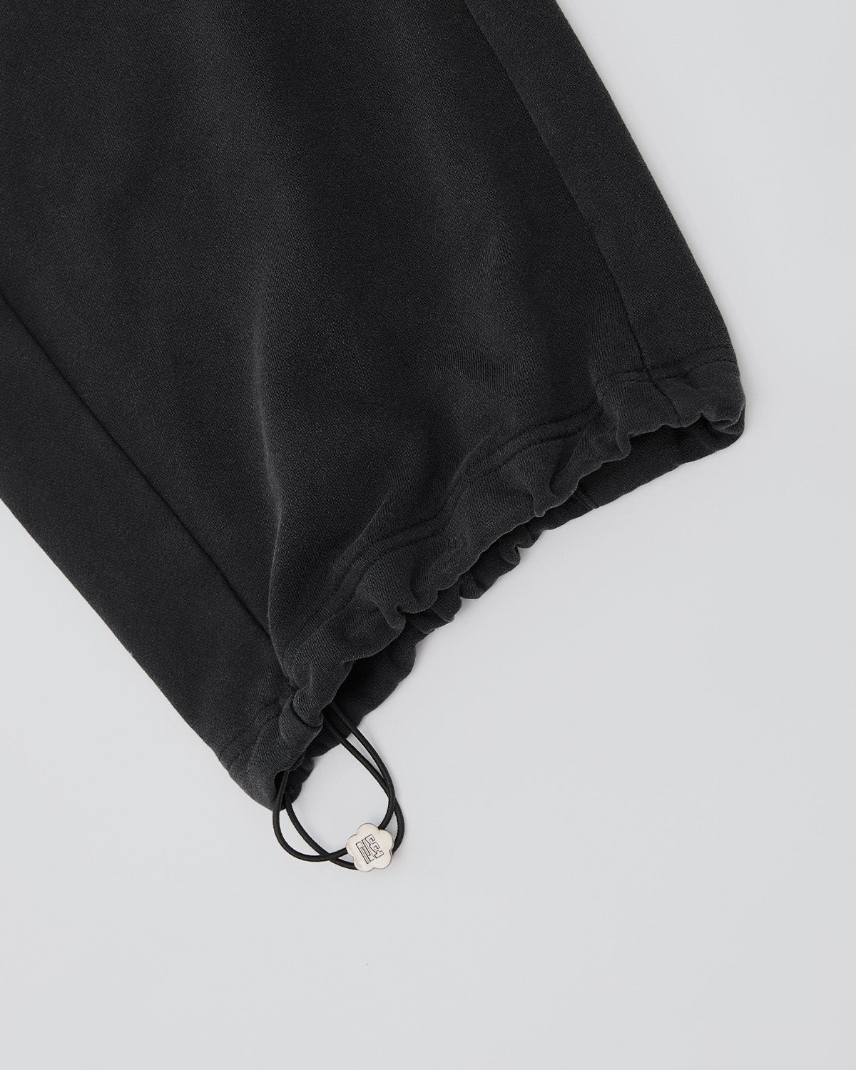 Close-up of Sundae School's FADE BLACK WRAP SWEATBAJI: wide-leg black cotton sweatpants with elasticated cuffs and an adjustable drawstring with metallic toggle, laid flat on a light gray background.
