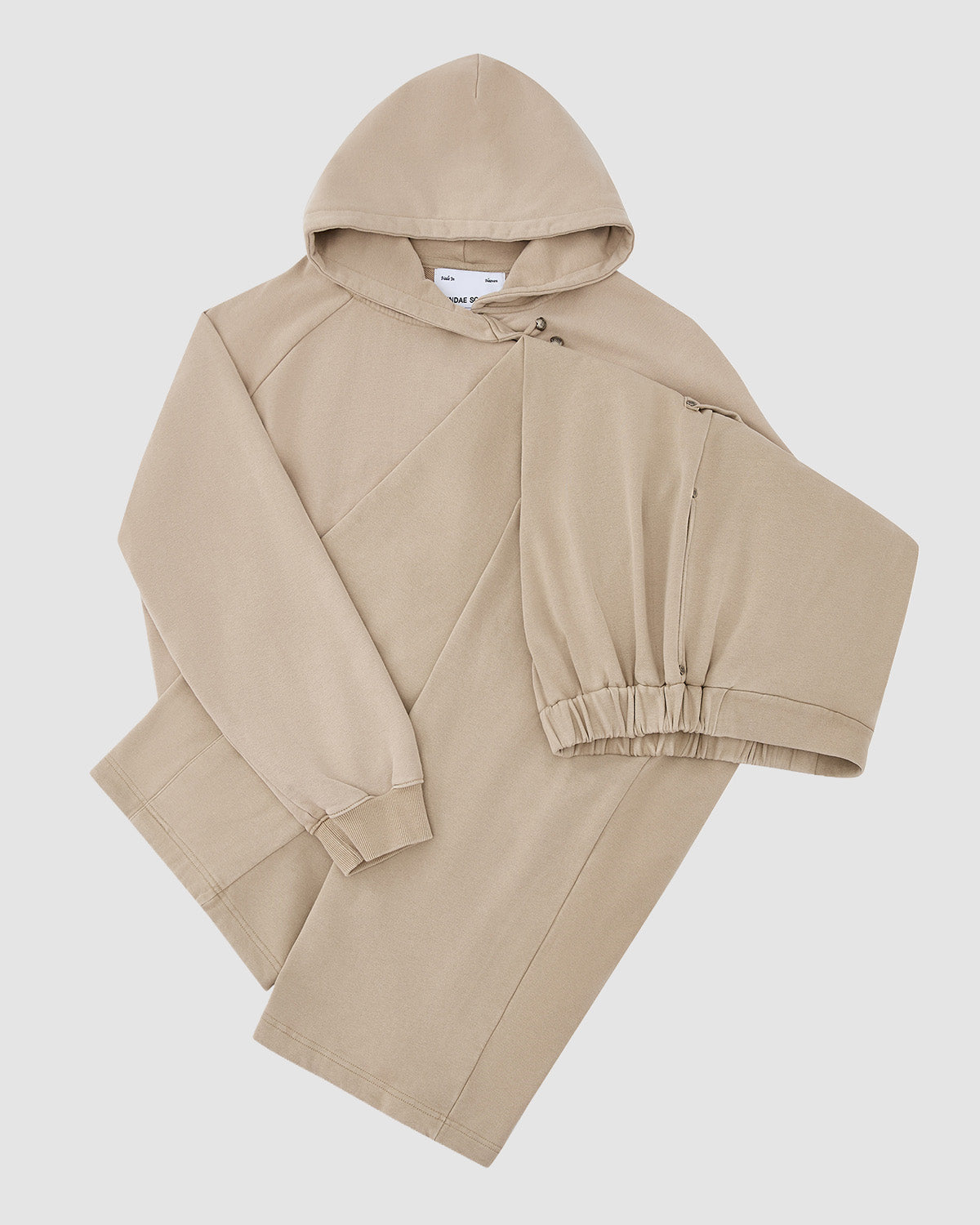 The TAUPE HANBOK HOODIE by Sundae School features a beige "떨" logo, diagonal seam design, long sleeves, one elastic cuff, and a signature sleeve stash pocket, shown laid flat on a white background.