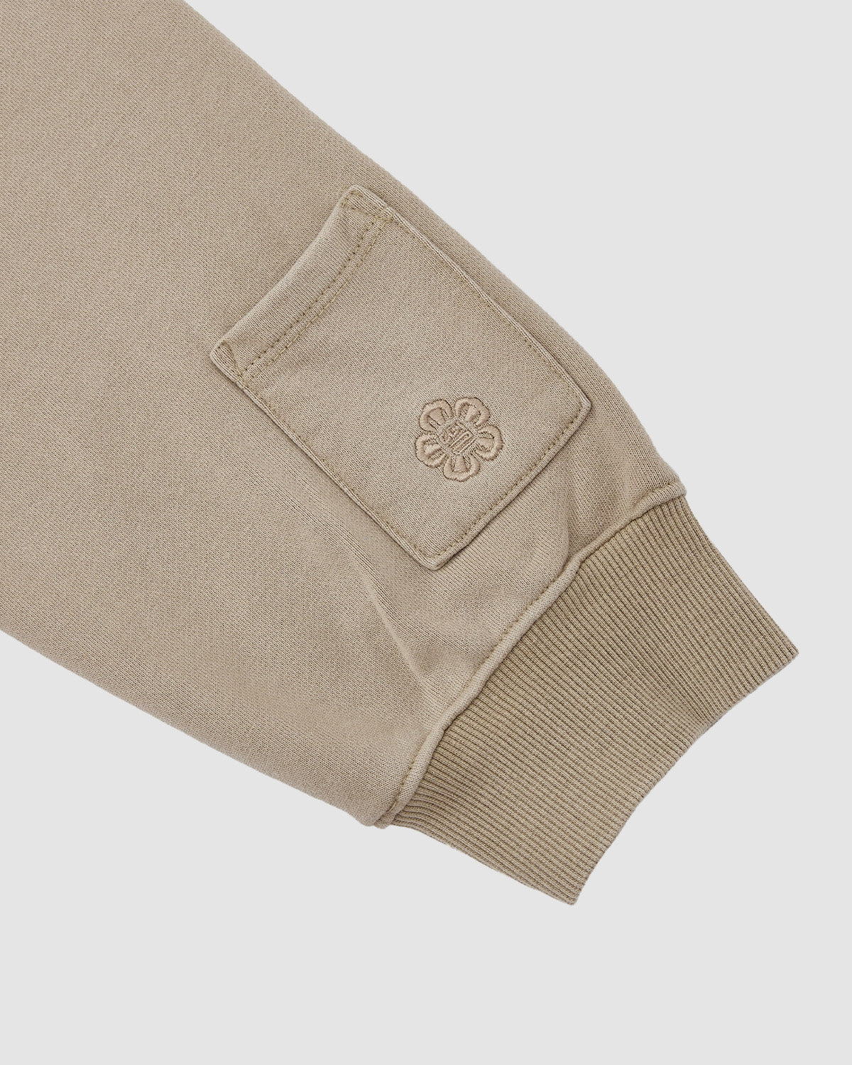 The TAUPE HANBOK HOODIE by Sundae School features beige long sleeves with ribbed knit cuffs and a signature rectangular sleeve stash pocket above the cuff, adorned with an embroidered floral or geometric emblem.