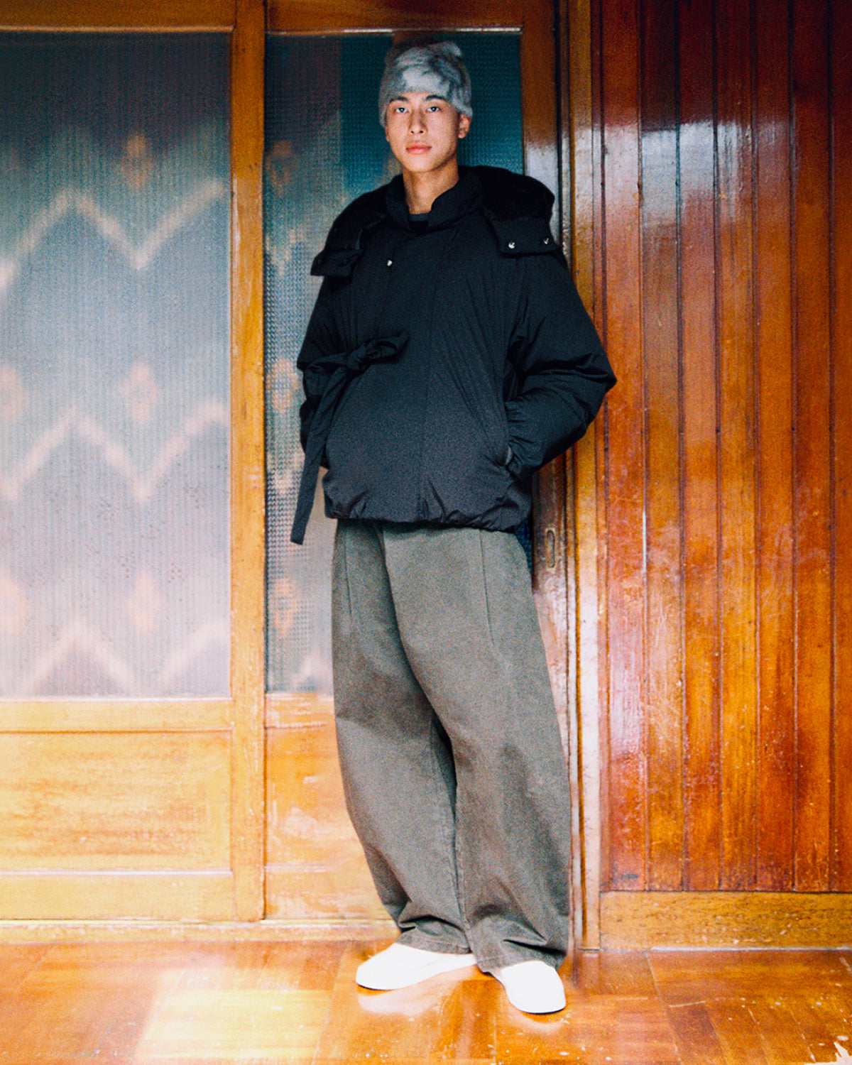 A person wearing the Sundae School BLACK IBUL DOWN PUFFER, loose dark pants, white shoes, and a light gray beanie stands indoors in front of wooden sliding doors.