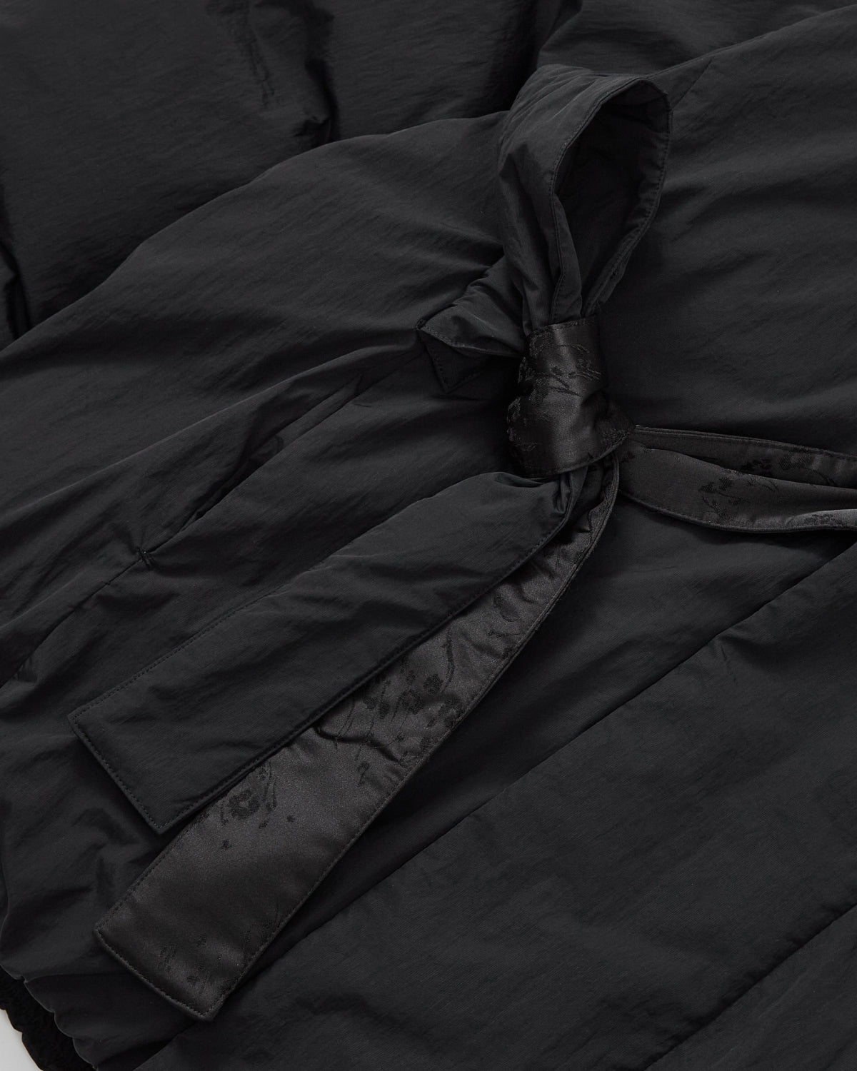 Close-up of the BLACK IBUL DOWN PUFFER by Sundae School, featuring black fabric with a matching belt tied in a knot. The slightly shiny material showcases gentle folds and creases, evoking the signature cocoon puffer look.