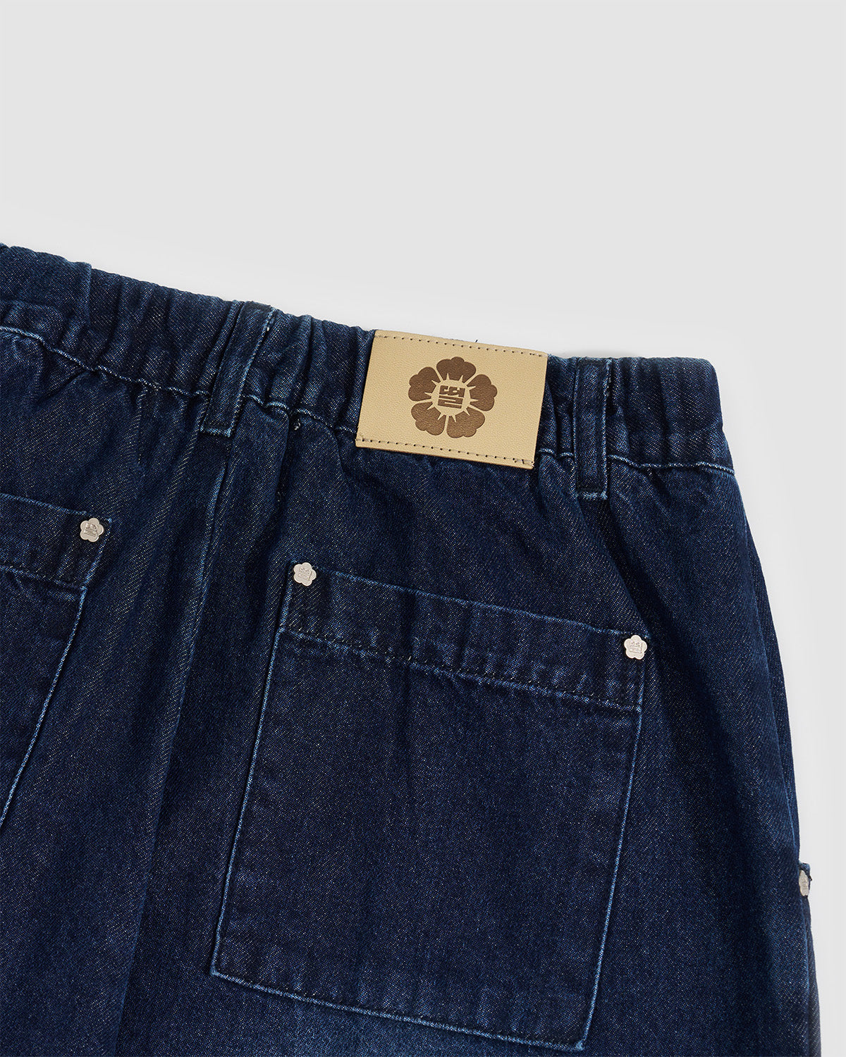 Close-up detail of Sundae School tan leather brand patch with embossed flower logo on indigo fade hanbok denim waistband, showing back pocket construction with silver snap button hardware