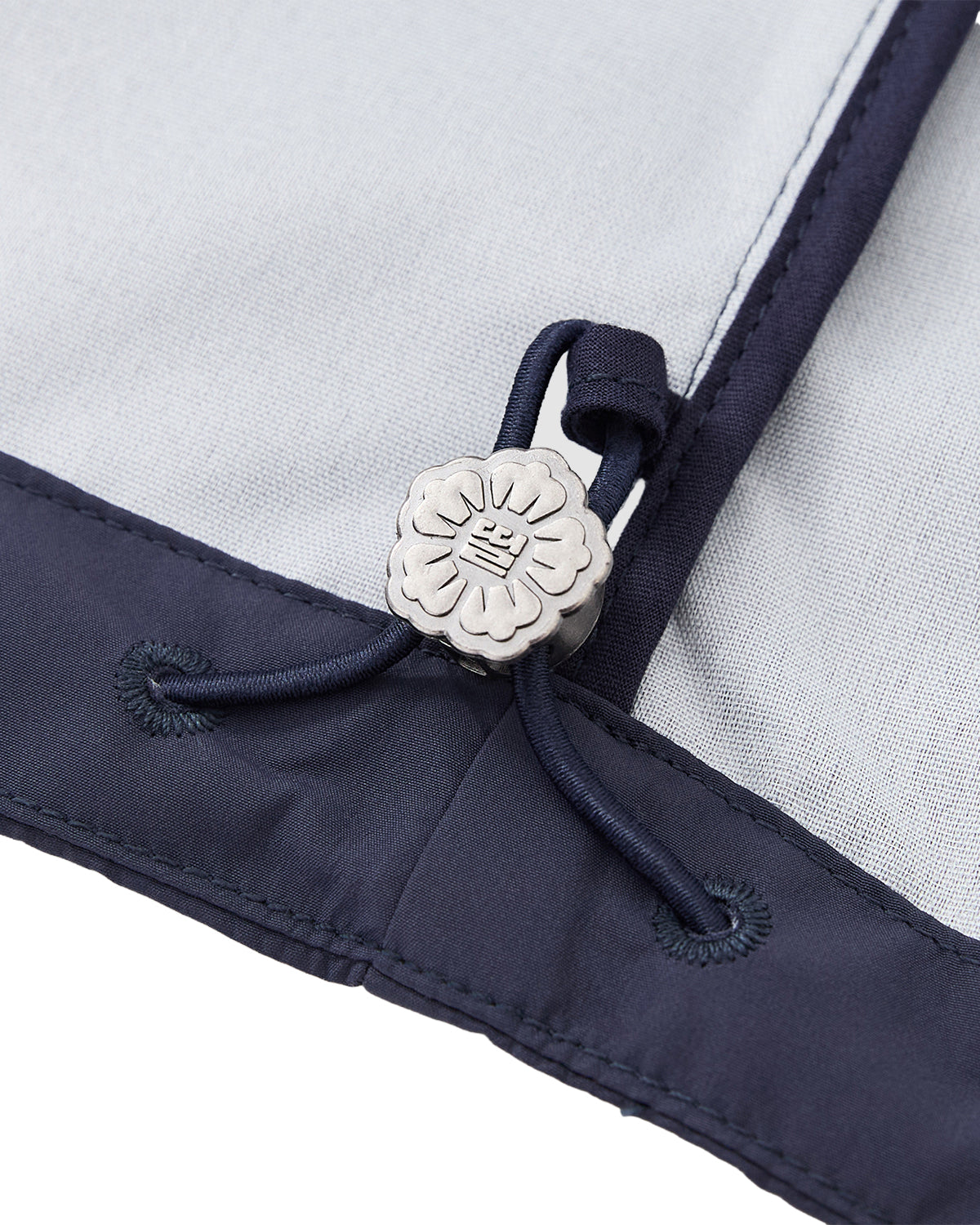 Detailed macro shot of an adjustable drawstring hem on a navy blue shell jacket, featuring a custom silver flower-shaped cord stopper engraved with a traditional emblem.