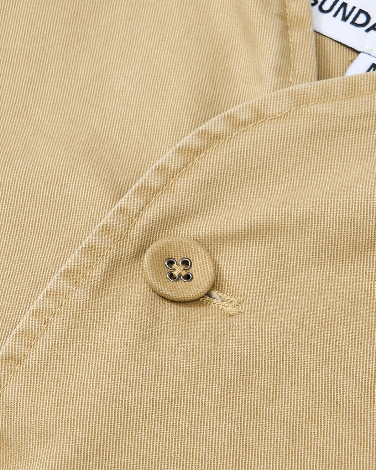 Close-up of the TAN JEOGORI TIE JACKET by Sundae School, featuring beige fabric with a sewn button, buttonhole detail, part of the collar, and a partially visible tag with text.